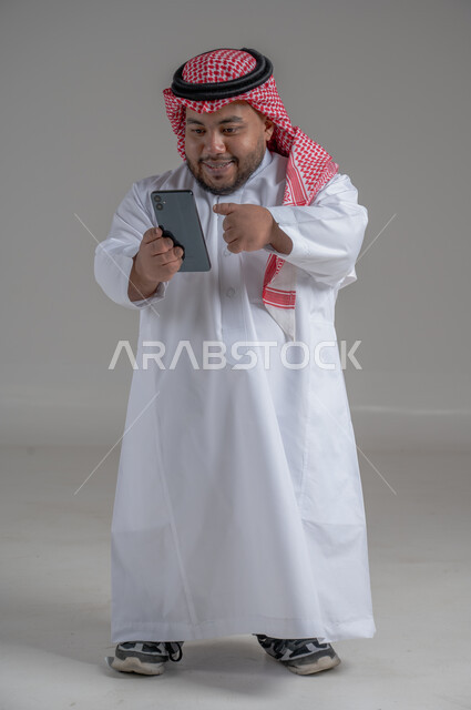 إجراء مكالمات الفيديو مع الأهل والأصدقاء، استخدام الجوال في أخذ لقطات السيلفي، بورتريه لشاب وسيم عربي خليجي سعودي قصير القامة  يرتدي الثوب التقليدي والشماغ يمسك الهاتف المحمول بيده وينظر إليه بإيماءات السعادة، الجسم بالطول الكامل، خلفية رمادية