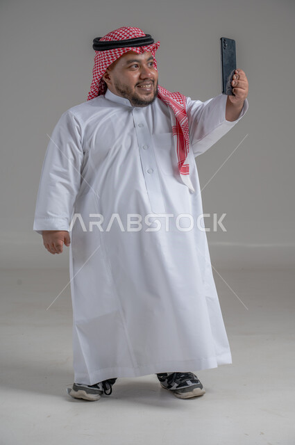 إجراء مكالمات الفيديو مع الأهل والأصدقاء، استخدام الجوال في أخذ لقطات السيلفي، بورتريه لشاب وسيم عربي خليجي سعودي قصير القامة  يرتدي الثوب التقليدي والشماغ يمسك الهاتف المحمول بيده وينظر إليه بإيماءات السعادة، الجسم بالطول الكامل، خلفية رمادية