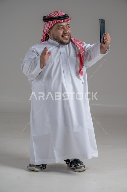 إجراء مكالمات الفيديو مع الأهل والأصدقاء، استخدام الجوال في أخذ لقطات السيلفي، بورتريه لشاب وسيم عربي خليجي سعودي قصير القامة  يرتدي الثوب التقليدي والشماغ يمسك الهاتف المحمول بيده وينظر إليه بإيماءات السعادة، الجسم بالطول الكامل، خلفية رمادية