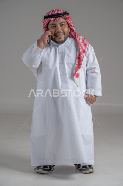 Communicating and chatting with family and friends, following the latest developments around the world, integrating modern techniques and technology into practical life, portrait of a handsome young Saudi Arabian Gulf man, short in stature, wearing a traditional thobe and a shemagh, holding a mobile phone in his hand, full body, gray background