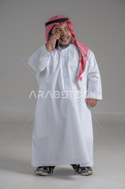 التواصل والدردشة مع الأهل والأصدقاء, متابعة آخر المستجدات حول العالم, دمج التقنيات الحديثة والتكنولوجيا بالحياة العملية, بورتريه لشاب وسيم عربي خليجي سعودي قصير القامة يرتدي الثوب التقليدي والشماغ  يمسك الهاتف المحمول بيده، الجسم بالطول الكامل، خلفية رمادية