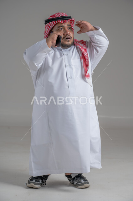 Communicating and chatting with family and friends, following the latest developments around the world, integrating modern techniques and technology into practical life, portrait of a handsome young Saudi Arabian Gulf man, short in stature, wearing a traditional thobe and a shemagh, holding a mobile phone in his hand, full body, gray background