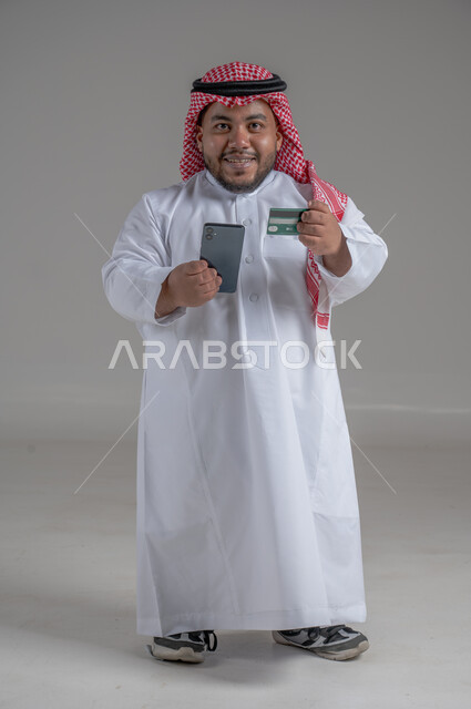 Concept of saving time and effort, portrait of a short Saudi Arabian young man wearing traditional thobe and shemagh using smartphone and credit card for online shopping, use of modern and advanced technical devices to meet needs, full body image, gray background
