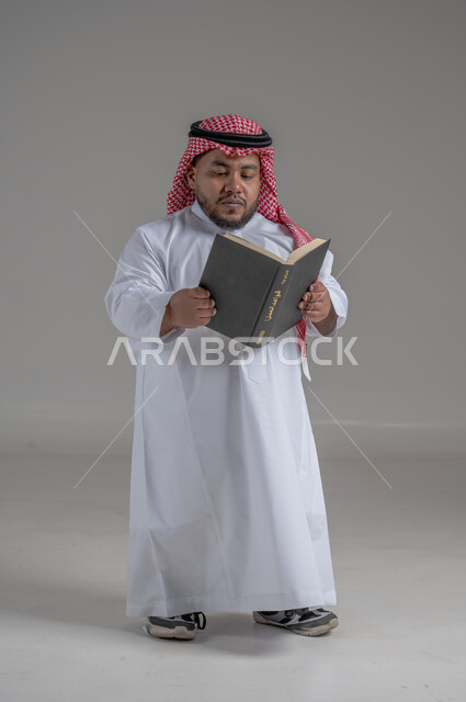 مفهوم التثقيف والبحث العلمي، هواية المطالعة والاستكشاف، قضاء وقت الفراغ بالتعلم والثقافة، بورتريه لرجل عربي خليجي سعودي قصير القامة مبتسم يرتدي الزي التقليدي والشماغ  يحمل بيده كتاب, صورة الجسم بالطول الكامل، خلفية رمادية