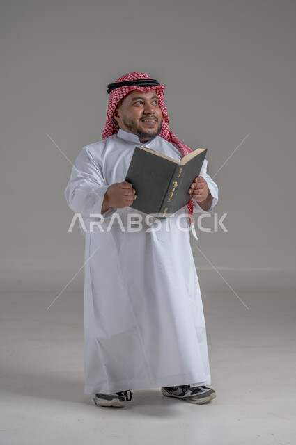 مفهوم التثقيف والبحث العلمي، هواية المطالعة والاستكشاف، قضاء وقت الفراغ بالتعلم والثقافة، بورتريه لرجل عربي خليجي سعودي قصير القامة مبتسم يرتدي الزي التقليدي والشماغ  يحمل بيده كتاب, صورة الجسم بالطول الكامل، خلفية رمادية