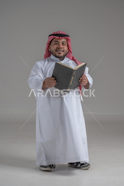 مفهوم التثقيف والبحث العلمي، هواية المطالعة والاستكشاف، قضاء وقت الفراغ بالتعلم والثقافة، بورتريه لرجل عربي خليجي سعودي قصير القامة مبتسم يرتدي الزي التقليدي والشماغ  يحمل بيده كتاب, صورة الجسم بالطول الكامل، خلفية رمادية