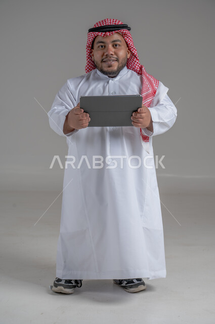 استخدام التكنولوجيا و التقنيات الحديثة، بورتريه لرجل عربي خليجي سعودي قصير القامة يرتدي الشماغ و الثوب التقليدي يحمل بيده جهاز التابليت، الدردشة مع الأهل والأصدقاء, تصفح برامج التواصل الاجتماعي، تطبيقات وبرامج الأجهزة اللوحية، صورة الجسم بالطول الكامل، خلفية رمادية