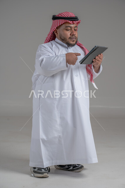 استخدام التكنولوجيا و التقنيات الحديثة، بورتريه لرجل عربي خليجي سعودي قصير القامة يرتدي الشماغ و الثوب التقليدي يحمل بيده جهاز التابليت، الدردشة مع الأهل والأصدقاء, تصفح برامج التواصل الاجتماعي، تطبيقات وبرامج الأجهزة اللوحية، صورة الجسم بالطول الكامل، خلفية رمادية
