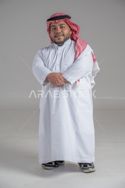 التمكين الاجتماعي لذوي الهمم، بورتريه لشاب عربي خليجي سعودي قصير القامة يرتدي الثوب التقليدي والشماغ يقوم بتحريك يديه بايماءات تدل على السعادة والسرور، صورة الجسم بالطول الكامل، خلفية رمادية