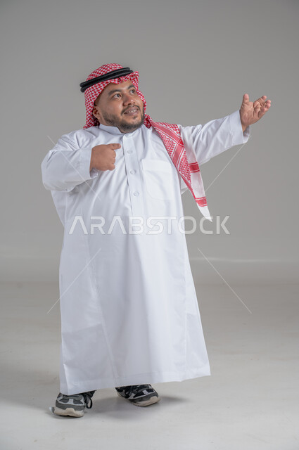 التمكين الاجتماعي لذوي الهمم، بورتريه لشاب عربي خليجي سعودي قصير القامة يرتدي الثوب التقليدي والشماغ يقوم بتحريك يديه بايماءات تدل على السعادة والسرور، صورة الجسم بالطول الكامل، خلفية رمادية