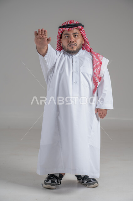 التمكين الاجتماعي لذوي الهمم، بورتريه لشاب عربي خليجي سعودي قصير القامة يرتدي الثوب التقليدي والشماغ يقوم بتحريك يديه بايماءات تدل على السعادة والسرور، صورة الجسم بالطول الكامل، خلفية رمادية