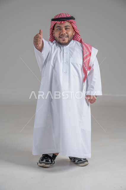 التمكين الاجتماعي لذوي الهمم، بورتريه لشاب عربي خليجي سعودي قصير القامة يرتدي الثوب التقليدي والشماغ يقوم بتحريك يديه بايماءات تدل على السعادة والسرور، صورة الجسم بالطول الكامل، خلفية رمادية