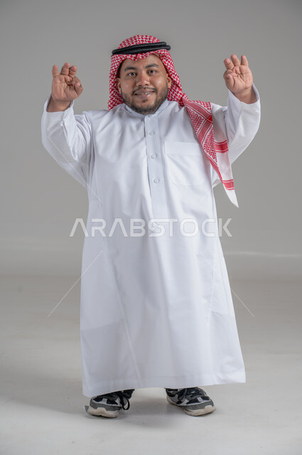 التمكين الاجتماعي لذوي الهمم، بورتريه لشاب عربي خليجي سعودي قصير القامة يرتدي الثوب التقليدي والشماغ يقوم بتحريك يديه بايماءات تدل على السعادة والسرور، صورة الجسم بالطول الكامل، خلفية رمادية