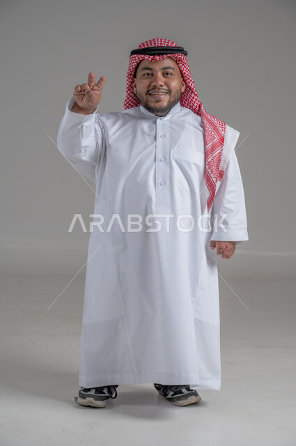 مفهوم الفوز والانتصار، بورتريه لشاب عربي خليجي سعودي قصير القامة يرتدي الثوب التقليدي والشماغ يقوم بتحريك يديه بايماءات تدل على السعادة والسرور، صورة الجسم بالطول الكامل، خلفية رمادية