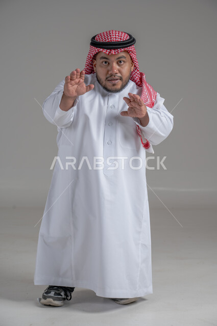 التمكين الاجتماعي لذوي الهمم، بورتريه لشاب عربي خليجي سعودي قصير القامة يرتدي الثوب التقليدي والشماغ يقوم بتحريك يديه بايماءات تدل على السعادة والسرور، صورة الجسم بالطول الكامل، خلفية رمادية