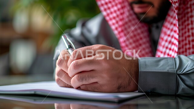 مهن ووظائف ادارية مكتبية، بيئة العمل المريحة في الشركات السعودية، صورة مقربة لرجل اعمال عربي خليجي سعودي يرتدي الثوب التقليدي والشماغ يجلس داخل المكتب في مقر الشركة يقوم بتدوين وتسجيل الملاحظات