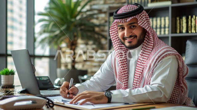 بيئة العمل المريحة في الشركات السعودية، صورة مقربة لرجل اعمال عربي خليجي سعودي يرتدي الثوب التقليدي والشماغ يجلس داخل المكتب في مقر الشركة يقوم بتدوين وتسجيل الملاحظات، النظر الى الكاميرا بتعابير الفرح والسرور، مهن ووظائف ادارية مكتبية