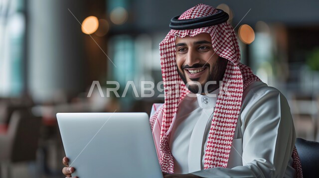 دمج التقنية والتكنولوجيا مع الحياة العملية, بيئة العمل المريحة في الشركات السعودية, صورة مقربة لرجل عربي خليجي سعودي مبتسم يرتدي الشماغ والثوب التقليدي يقوم بالعمل على الحاسوب المحمول بإيماءات السرور والفرح,  الجلوس على المكتب في مقر الشركة, إدارة وتنظيم الأعمال