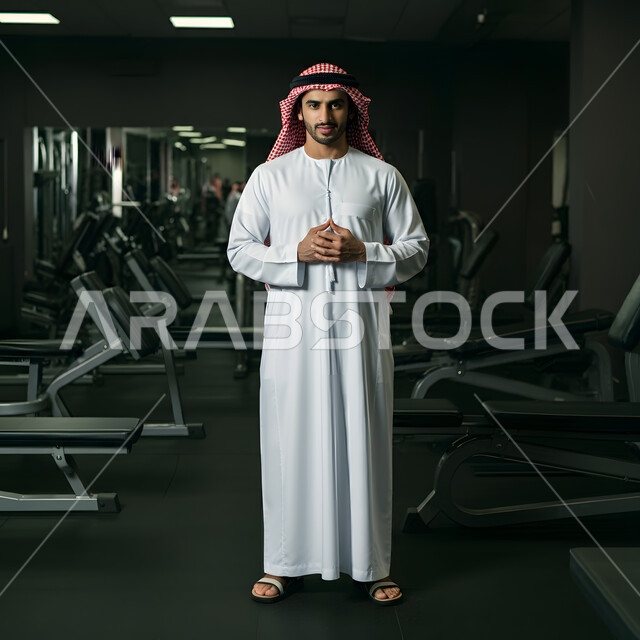 مفهوم الحفاظ على اللياقة، أدوات ومعدات رياضية، نشاط بدني للعناية بالصحة الجسدية، صورة مقربة لرجل عربي خليجي سعودي يرتدي الزي التقليدي والشماغ يقف في صالة الرياضة بالمملكة العربية السعودية، نادي رياضي رجالي، الجسم الكامل بالطول
