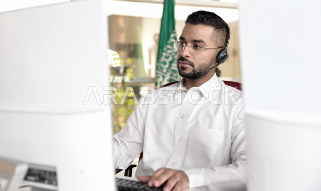موظف عربي خليجي سعودي يعمل في خدمة العملاء ويرتدي سماعة الاتصال، استعلامات واستفسارات، الاجابه على اسئلة العملاء، اجراء اتصال فيديو من خلال الحاسوب، نظارة طبية، متابعة وتطوير العمل