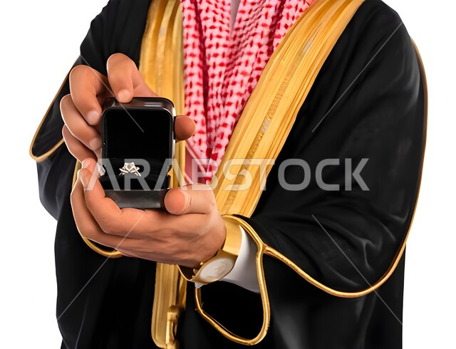 مراسم الاعراس والاحتفالات في المملكة، صورة مقربة ليد عريس عربي خليجي سعودي يرتدي الزي التقليدي والبشت الاسود يحمل بيده خاتم مرصع بالجواهر، مفهوم المناسبات والأفراح، أصالة العادات والتقاليد، خلفية بيضاء