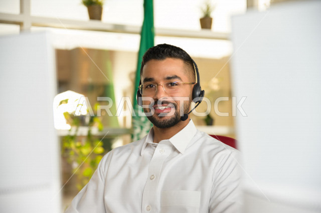 موظف عربي خليجي سعودي يعمل في خدمة العملاء ويرتدي سماعة الاتصال، استعلامات واستفسارات، الاجابه على اسئلة العملاء، اجراء اتصال فيديو من خلال الحاسوب، نظارة طبية، متابعة وتطوير العمل