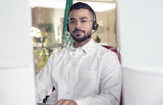 موظف عربي خليجي سعودي يعمل في خدمة العملاء ويرتدي سماعة الاتصال، استعلامات واستفسارات، الاجابه على اسئلة العملاء، اجراء اتصال فيديو من خلال الحاسوب، نظارة طبية، متابعة وتطوير العمل