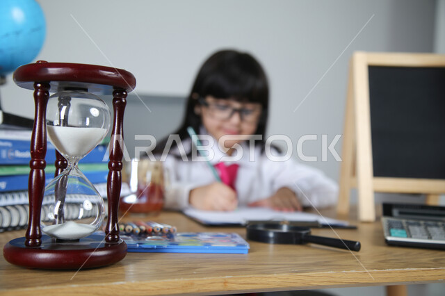 تجربة الأطفال للعمل في مجال التعليم، اختيار مهنة المعلم منذ الصغر، السعي نحو تحقيق الطموحات المستقبلية، بورتريه مقرب لفتاة عربية خليجية سعودية ترتدي الزي الرسمي والنظارات الطبية تجلس خلف المكتب تمسك القلم وتصحح الأوراق، الساعة الرملية والقرطاسية، خلفية رمادية