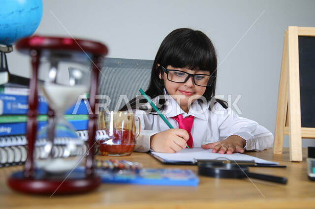 تجربة الأطفال للعمل في مجال التعليم، اختيار مهنة المعلم منذ الصغر، السعي نحو تحقيق الطموحات المستقبلية، بورتريه مقرب لفتاة عربية خليجية سعودية ترتدي الزي الرسمي والنظارات الطبية تجلس خلف المكتب تمسك القلم وتصحح الأوراق، الساعة الرملية والقرطاسية، خلفية رمادية