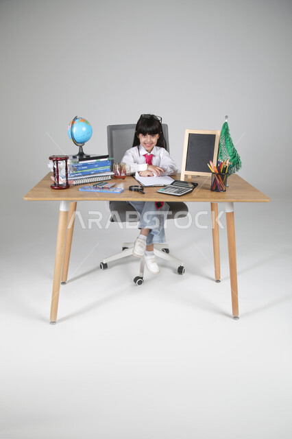 تجربة الأطفال للعمل في مجال التعليم منذ الصغر، السعي نحو تحقيق الطموحات المستقبلية، بورتريه مقرب لفتاة عربية خليجية سعودية ترتدي الزي الرسمي للمعلم تجلس خلف المكتب تنظر للكاميرا بإيماءات السعادة، الساعة الرملية والقرطاسية، الجسم بالطول الكامل، خلفية رمادية