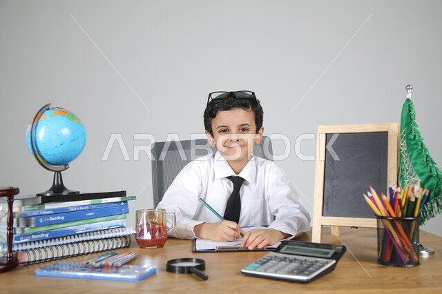 اختيار مهنة المعلم منذ الصغر، السعي نحو تحقيق الطموحات والأحلام المستقبلية، تجربة العمل في مجال التعليم، بورتريه مقرب لولد عربي خليجي سعودي يرتدي الزي الرسمي يجلس خلف المكتب وينظر للكاميرا بإيماءات السرور، المجسمات التعليمية والعلم والقرطاسية، خلفية رمادية