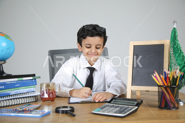 تجربة العمل في مجال التعليم، اختيار مهنة المعلم منذ الصغر، السعي نحو تحقيق الطموحات والأحلام المستقبلية، بورتريه مقرب لولد عربي خليجي سعودي يرتدي الزي الرسمي يجلس خلف المكتب يمسك القلم ويقوم بتصحيح الأوراق، المجسمات التعليمية والعلم والقرطاسية، خلفية رمادية