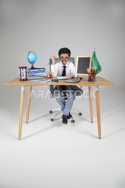 السعي نحو تحقيق الطموحات والأحلام المستقبلية، اختيار مهنة المعلم من الصغر، بورتريه لولد عربي خليجي سعودي يرتدي الزي الرسمي ويضع النظارة الطبية يجلس خلف المكتب وينظر للكاميرا بإيماءات السرور والثقة، المجسمات والعلم والقرطاسية، الجسم بالطول الكامل، خلفية رمادية