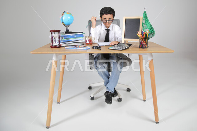 السعي نحو تحقيق الطموحات والأحلام المستقبلية، اختيار مهنة المعلم من الصغر، بورتريه لولد عربي خليجي سعودي يرتدي الزي الرسمي ويضع النظارة الطبية يجلس خلف المكتب وينظر للكاميرا بإيماءات السرور والثقة، المجسمات والعلم والقرطاسية، الجسم بالطول الكامل، خلفية رمادية