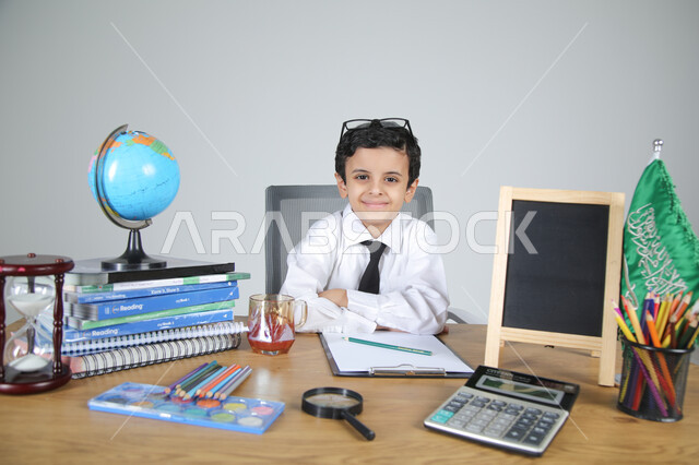 اختيار مهنة المعلم منذ الصغر، السعي نحو تحقيق الطموحات والأحلام المستقبلية، تجربة العمل في مجال التعليم، بورتريه مقرب لولد عربي خليجي سعودي يرتدي الزي الرسمي يجلس خلف المكتب وينظر للكاميرا بإيماءات السرور، المجسمات التعليمية والعلم والقرطاسية، خلفية رمادية