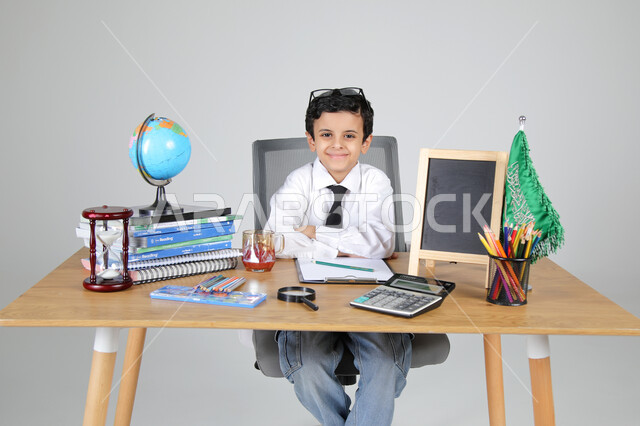 اختيار مهنة المعلم منذ الصغر، السعي نحو تحقيق الطموحات والأحلام المستقبلية، تجربة العمل في مجال التعليم، بورتريه مقرب لولد عربي خليجي سعودي يرتدي الزي الرسمي يجلس خلف المكتب وينظر للكاميرا بإيماءات السرور، المجسمات التعليمية والعلم والقرطاسية، خلفية رمادية