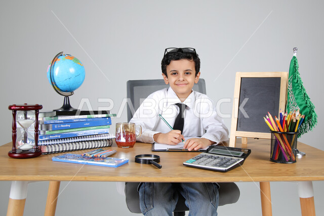 اختيار مهنة المعلم منذ الصغر، السعي نحو تحقيق الطموحات والأحلام المستقبلية، تجربة العمل في مجال التعليم، بورتريه مقرب لولد عربي خليجي سعودي يرتدي الزي الرسمي يجلس خلف المكتب وينظر للكاميرا بإيماءات السرور، المجسمات التعليمية والعلم والقرطاسية، خلفية رمادية