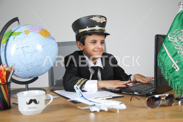 عيش التجربة والعمل في مجال الطيران باختيار مهنة الطيار، تجسيد المهن المستقبلية للأطفال، بورتريه مقرب لولد عربي خليجي سعودي يرتدي زي الطيار ويضع القبعة يجلس خلف المكتب ويعمل على الحاسوب المحمول بتركيز، العلم السعودي ومجسم الكرة الأرضية على الطاولة، خلفية رمادية