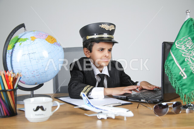 Experience and work in the aviation field by choosing the profession of a pilot, embodying future professions for children, close-up portrait of a Saudi Arabian Gulf boy wearing a pilot's uniform and hat sitting behind a desk and working on a laptop with concentration, Saudi flag and globe on the table, gray background