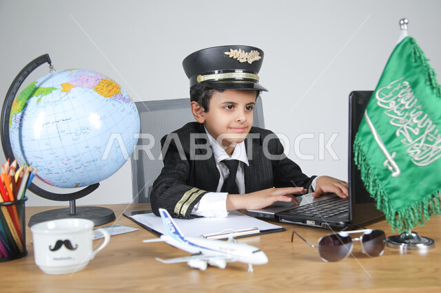 عيش التجربة والعمل في مجال الطيران باختيار مهنة الطيار، تجسيد المهن المستقبلية للأطفال، بورتريه مقرب لولد عربي خليجي سعودي يرتدي زي الطيار ويضع القبعة يجلس خلف المكتب ويعمل على الحاسوب المحمول بتركيز، العلم السعودي ومجسم الكرة الأرضية على الطاولة، خلفية رمادية