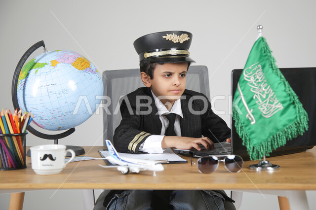 عيش التجربة والعمل في مجال الطيران باختيار مهنة الطيار، تجسيد المهن المستقبلية للأطفال، بورتريه مقرب لولد عربي خليجي سعودي يرتدي زي الطيار ويضع القبعة يجلس خلف المكتب ويعمل على الحاسوب المحمول بتركيز، العلم السعودي ومجسم الكرة الأرضية على الطاولة، خلفية رمادية