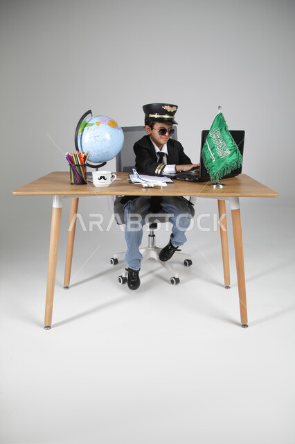 اختيار عيش تجربة كابتن الطيران، تجسيد المهن المستقبلية للأطفال، بورتريه لولد عربي خليجي سعودي يرتدي زي الطيار يضع القبعة النظارة الشمسية يجلس خلف المكتب ويعمل على الحاسوب المحمول، العلم السعودي ومجسم الكرة الأرضية على الطاولة، الجسم بالطول الكامل، خلفية رمادية