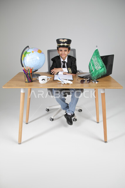 Choosing to live the experience of a flight captain, embodying future professions for children, portrait of a Saudi Arabian Gulf boy wearing a pilot's uniform and hat sitting behind a desk and looking at the camera with gestures of pleasure and confidence, Saudi flag and globe on the table, full-length body, gray background