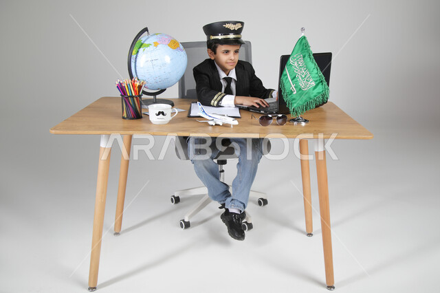 اختيار عيش تجربة كابتن الطيران، تجسيد المهن المستقبلية للأطفال، بورتريه لولد عربي خليجي سعودي يرتدي زي الطيار ويضع القبعة يجلس خلف المكتب ويعمل على الحاسوب المحمول بتركيز، العلم السعودي ومجسم الكرة الأرضية على الطاولة، الجسم بالطول الكامل، خلفية رمادية