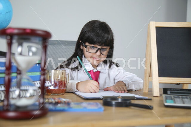 تجربة الأطفال للعمل في مجال التعليم، اختيار مهنة المعلم منذ الصغر، السعي نحو تحقيق الطموحات المستقبلية، بورتريه مقرب لفتاة عربية خليجية سعودية ترتدي الزي الرسمي والنظارات الطبية تجلس خلف المكتب تمسك القلم وتصحح الأوراق، الساعة الرملية والقرطاسية، خلفية رمادية