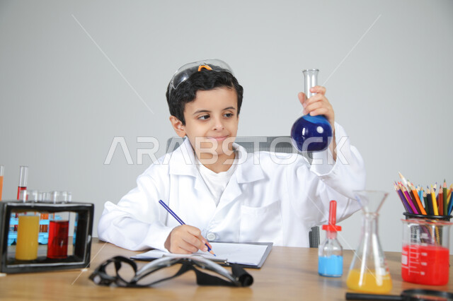 التجارب والأبحاث الكيميائية للعلماء الصغار، شغف الوصول إلى حلم دراسة الصيدلة مستقبلا، تجسيد مهن الأطفال المستقبلية منذ الصغر، بورتريه مقرب لصبي عربي خليجي سعودي يرتدي المعطف الطبي يجلس خلف المكتب ويمسك بزجاجات التحاليل ويقوم بتسجيل الملاحظات، خلفية رمادية