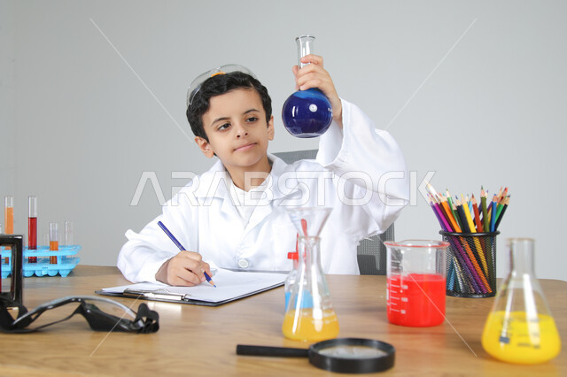 التجارب والأبحاث الكيميائية للعلماء الصغار، شغف الوصول إلى حلم دراسة الصيدلة مستقبلا، تجسيد مهن الأطفال المستقبلية منذ الصغر، بورتريه مقرب لصبي عربي خليجي سعودي يرتدي المعطف الطبي يجلس خلف المكتب ويمسك بزجاجات التحاليل ويقوم بتسجيل الملاحظات، خلفية رمادية