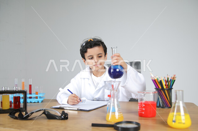 التجارب والأبحاث الكيميائية للعلماء الصغار، شغف الوصول إلى حلم دراسة الصيدلة مستقبلا، تجسيد مهن الأطفال المستقبلية منذ الصغر، بورتريه مقرب لصبي عربي خليجي سعودي يرتدي المعطف الطبي يجلس خلف المكتب ويمسك بزجاجات التحاليل ويقوم بتسجيل الملاحظات، خلفية رمادية