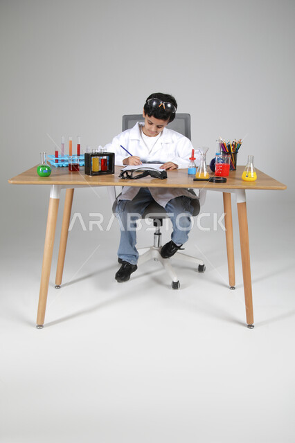 شغف الوصول إلى حلم دراسة الصيدلة مستقبلا، التجارب الكيميائية للعلماء الصغار، تجسيد مهن الأطفال المستقبلية منذ الصغر، بورتريه لصبي عربي خليجي سعودي يرتدي المعطف الطبي يجلس وأمامه زجاجات التحاليل ويقوم بتسجيل الملاحظات، الجسم بالطول الكامل، خلفية رمادية