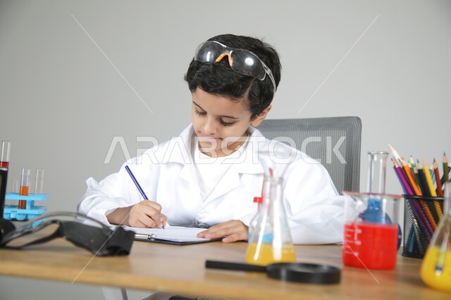 تجسيد مهن الأطفال المستقبلية منذ الصغر، شغف الوصول إلى حلم دراسة الصيدلة مستقبلا، التجارب الكيميائية للعلماء الصغار، بورتريه مقرب لصبي عربي خليجي سعودي يرتدي المعطف الطبي يجلس خلف المكتب وأمامه زجاجات التحاليل ويقوم بتسجيل الملاحظات بتركيز، خلفية رمادية